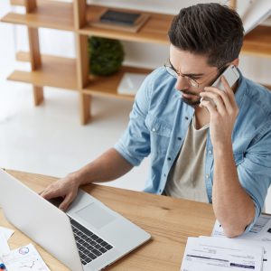 Bearded skilled young talented copywriter works on new article, keyboards at laptop, has phone conve